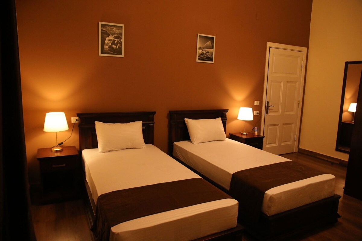 A hotel room with two beds, bedside lamps, a door, and two framed pictures on the wall.