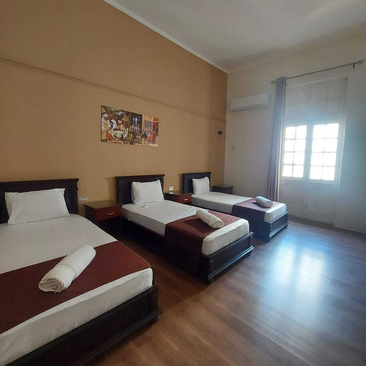 A hotel room with three beds, wooden flooring, a large window with curtains, and a painting on the wall.