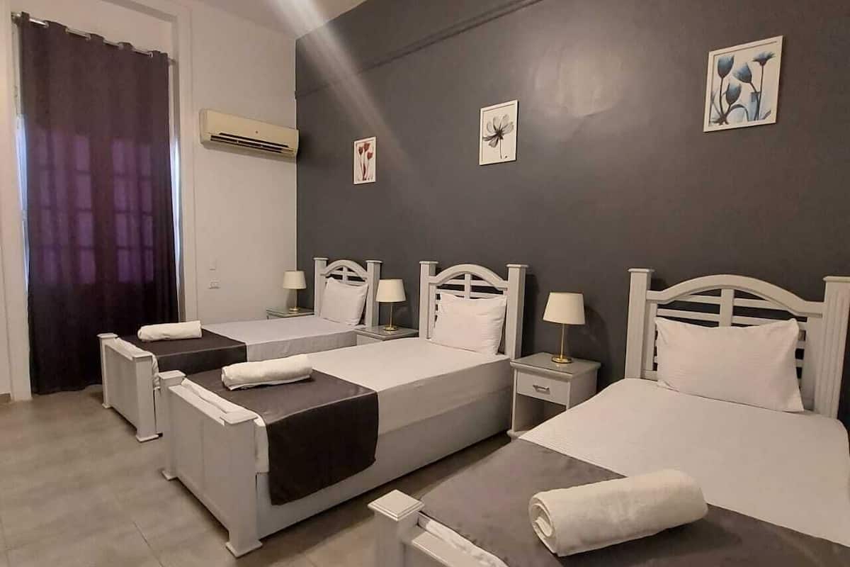 A hotel room with three single beds, a bathroom with a shower, and a large window with curtains.