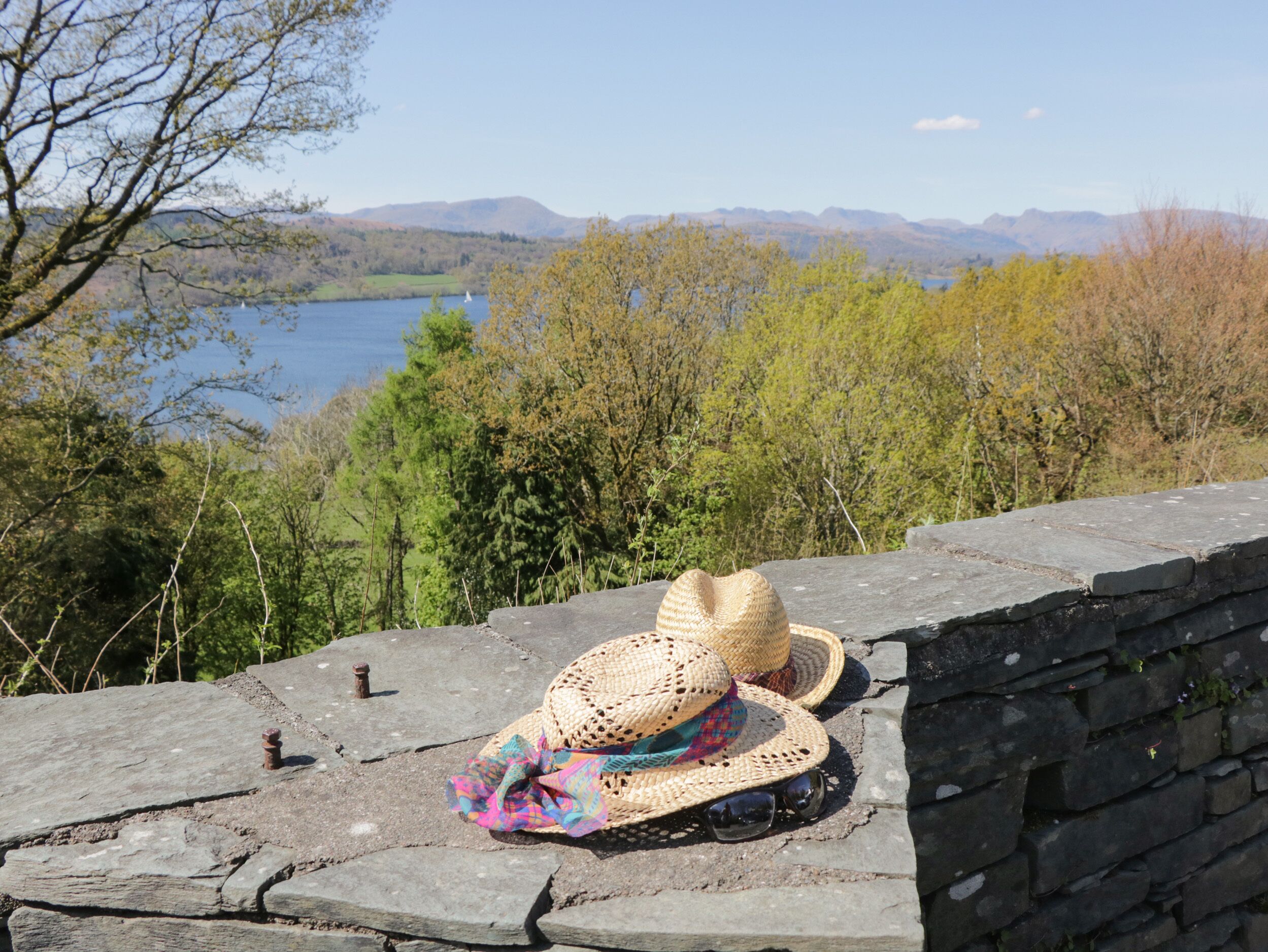 A stone wall with a straw hat and sunglasses placed on it, overlooking a lake and trees.