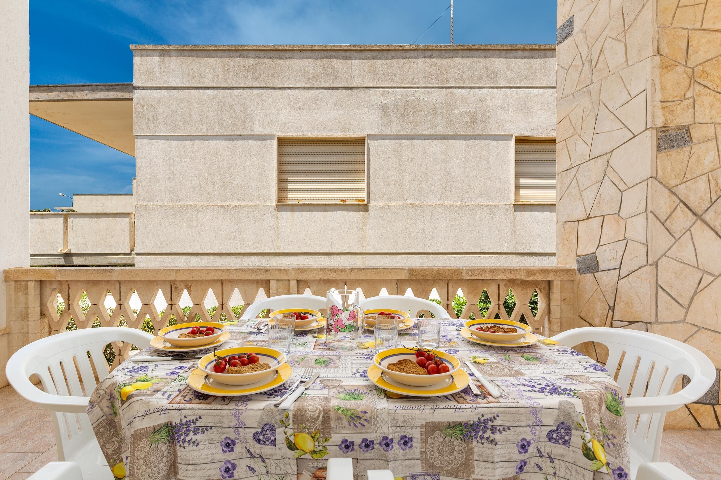 An outdoor dining area with a table set for four, surrounded by white chairs, against a building with stone walls and a clear blue sky.