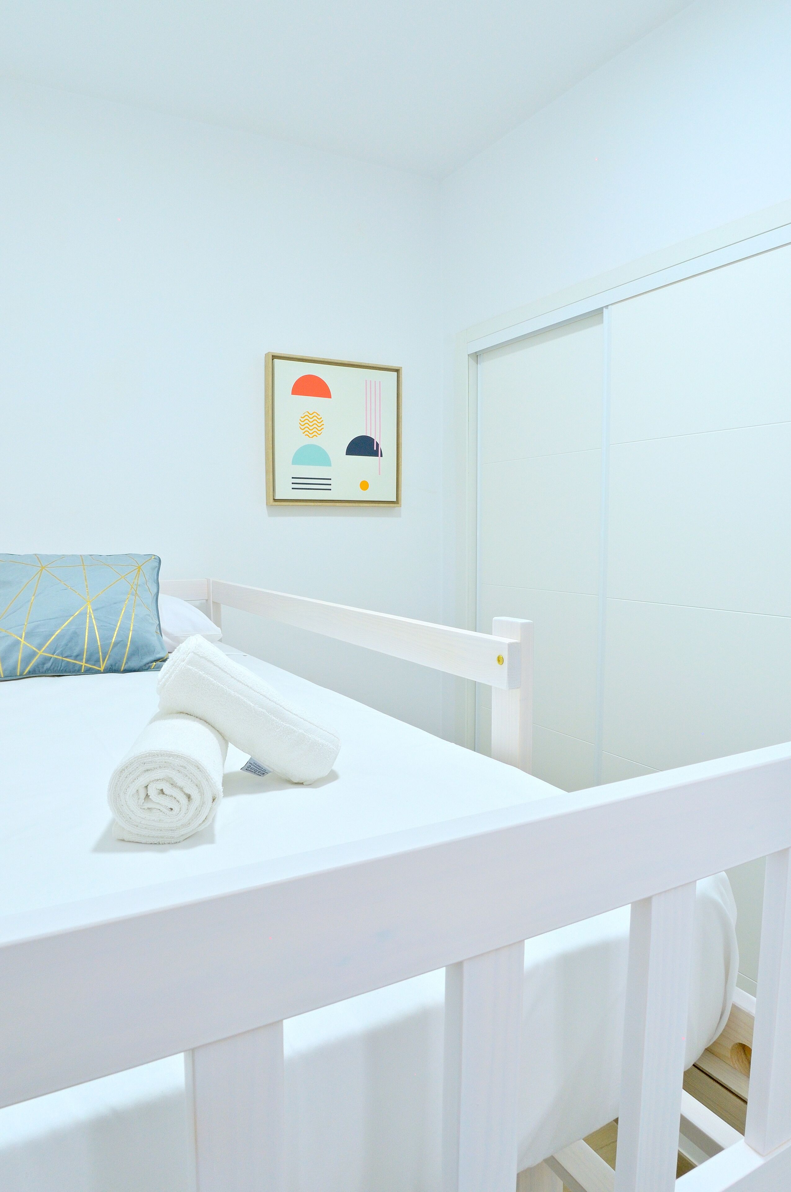 Un lit blanc avec une serviette blanche roulée, un coussin à motifs géométriques et un tableau encadré accroché au mur.