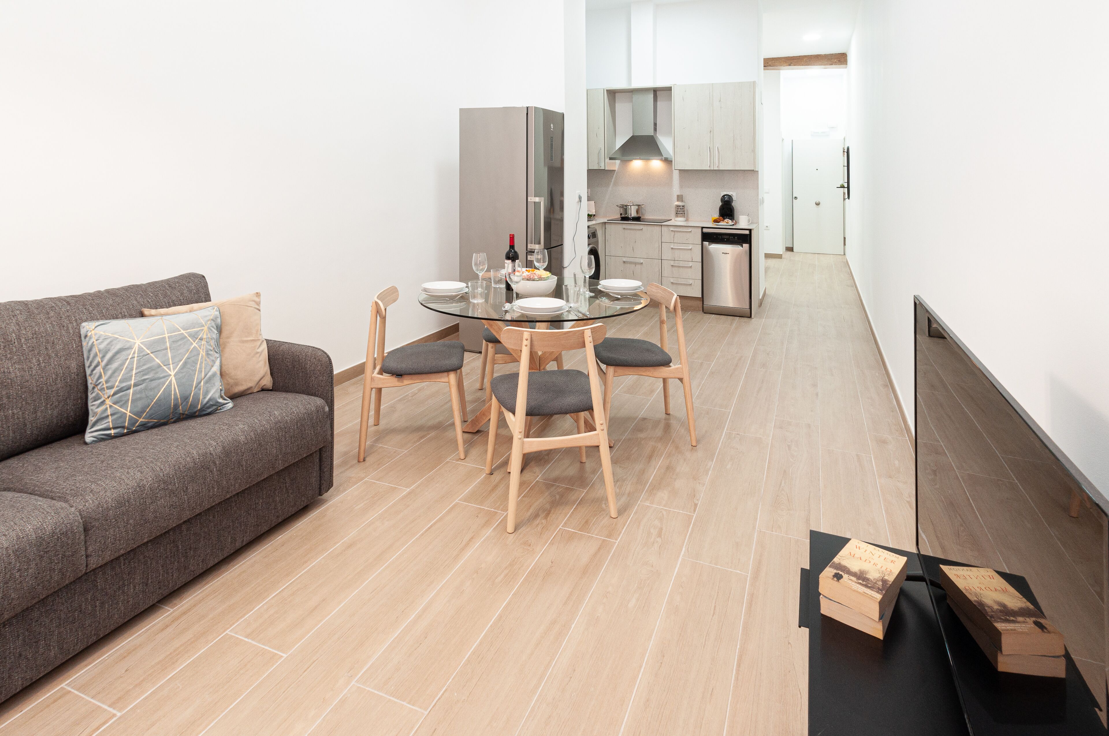 A modern living room with a grey sofa, a glass dining table with four chairs, and a kitchen in the background.