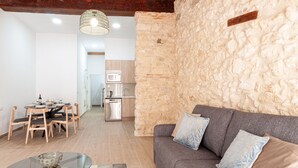 Een moderne woonkamer met een accentmuur van steen, een grijze bank met gekleurde kussens, een glazen salontafel en een eethoek met houten stoelen en een ronde tafel.
