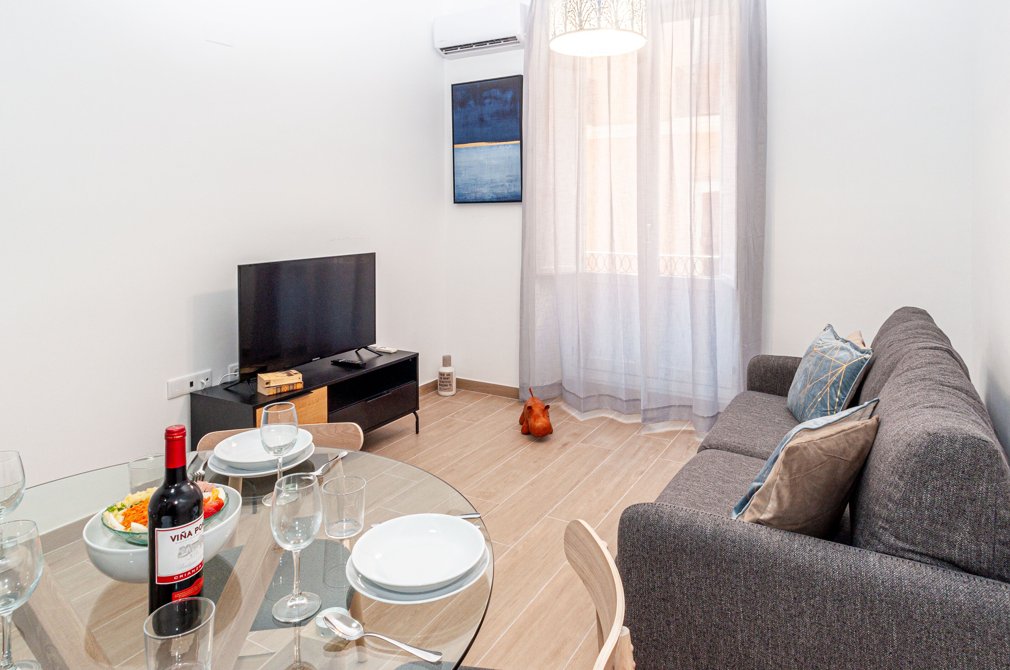 A modern living room with a round glass table, a sofa, a television, and a painting on the wall.