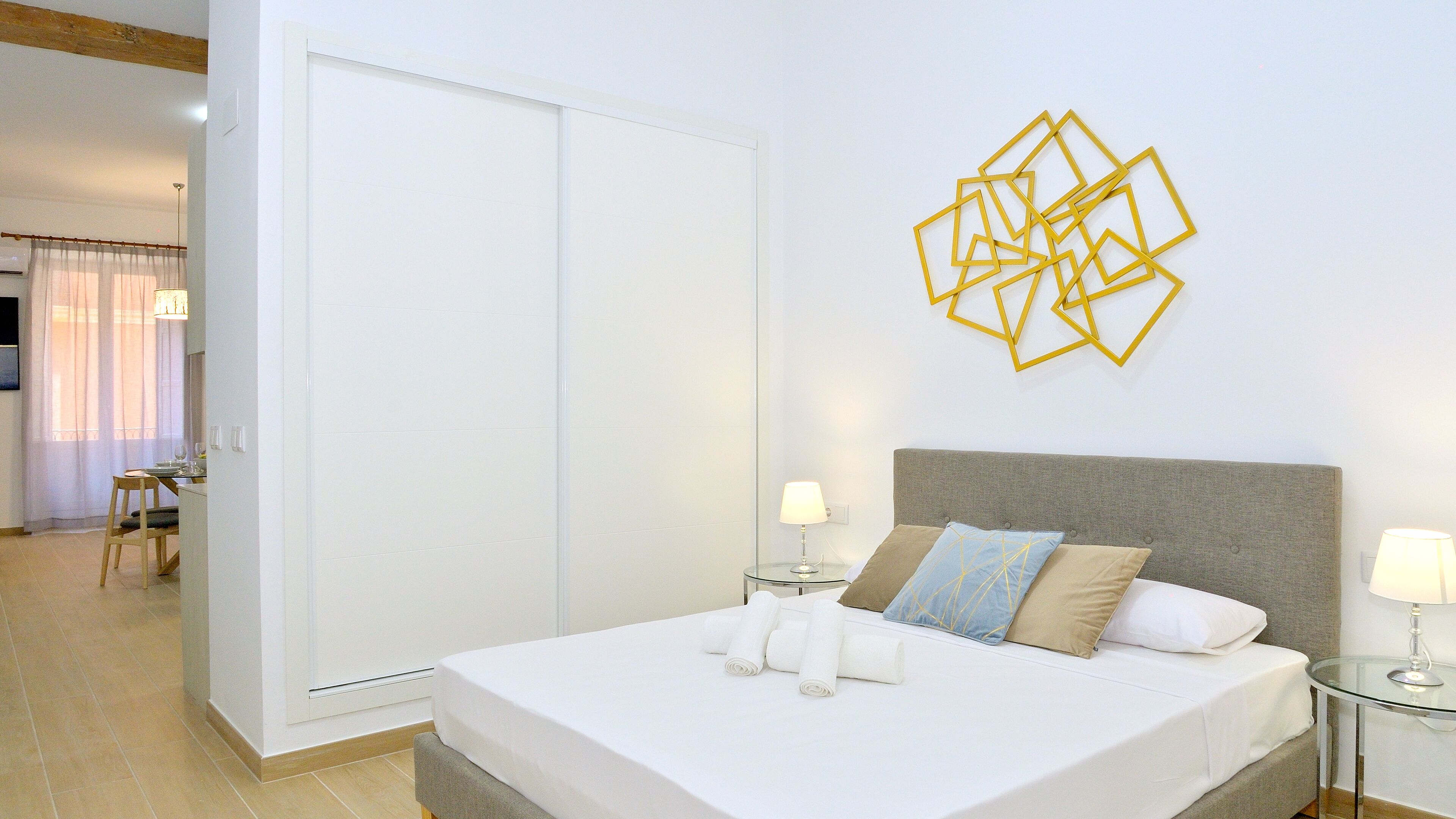 Moderni hotellihuone, jossa on suuri sänky, lasinen sivupöytä, yöpöytä ja geometrisesti kuvioitu seinäkoriste.