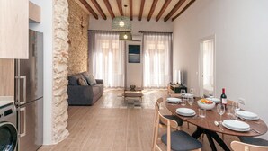 Een moderne keuken met een ingebouwde wasmachine, een eethoek met een houten tafel gedekt voor vier personen, en een zithoek met een bank en een stenen muur.
