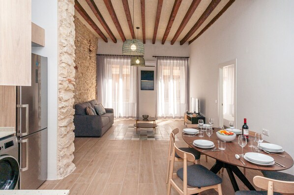 Une cuisine moderne équipée d’un lave-linge intégré, un coin repas avec une table en bois pour quatre personnes, et un salon avec un canapé et un mur en pierre.