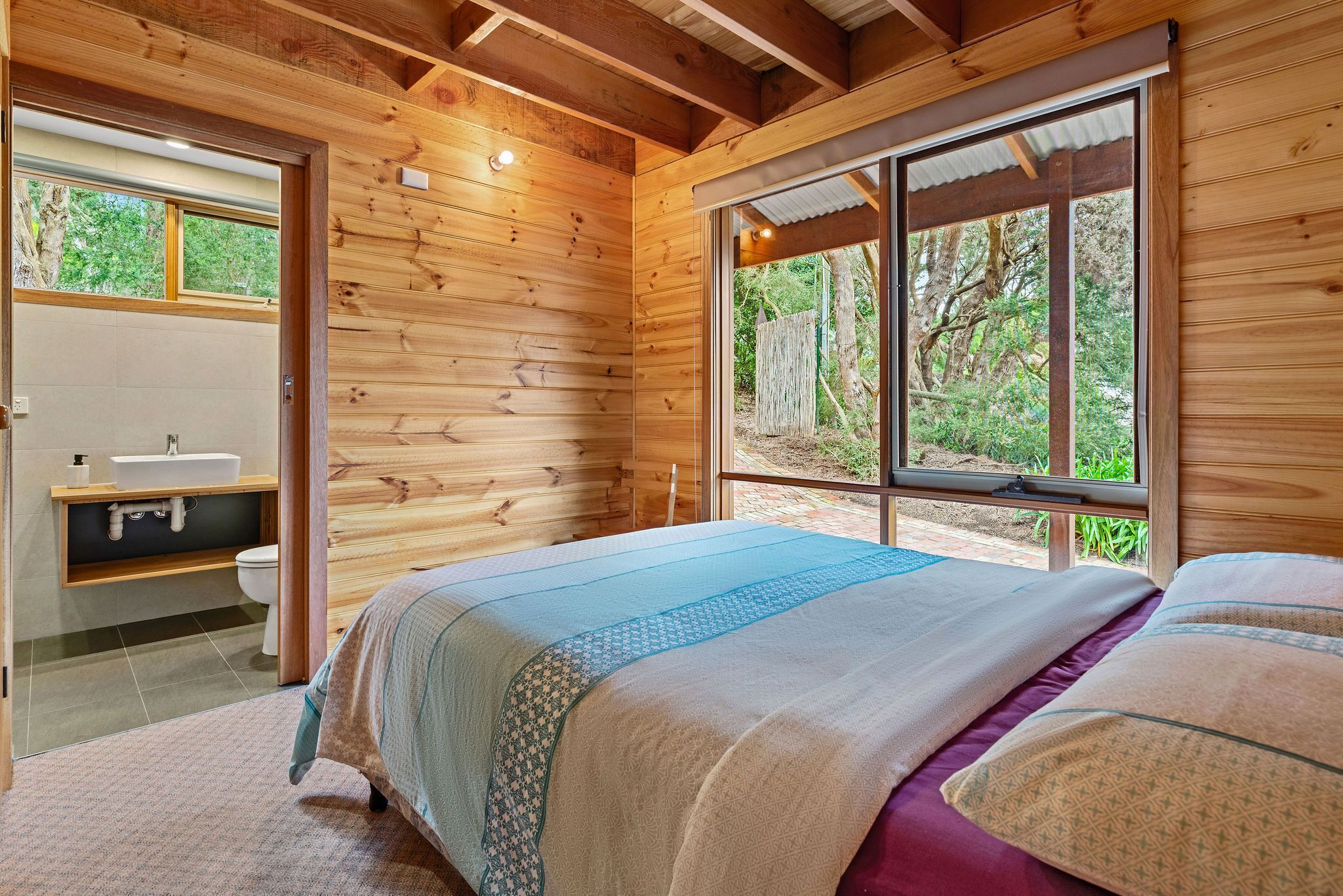 A wooden cabin bedroom with a bed, a window, and a bathroom with a sink and toilet.