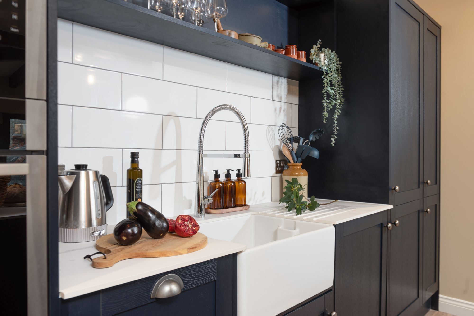 A modern kitchen with white tiles, a stainless steel kettle, a wooden cutting board with vegetables, and a black cabinet with a plant.