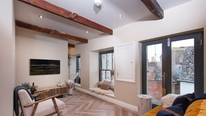 A spacious living room with a herringbone floor, wooden beams, a sofa with colorful cushions, a wooden bench, and a large window offering a view of the city.