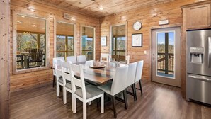 A wooden cabin interior with a dining table, chairs, a refrigerator, and large windows offering views of the outdoors. - Smokey Treats 5 Bedroom Cabin by RedAwning (Gatlinburg)