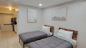 Quarto com duas camas, cabeceira de madeira e um corredor pequeno que dá para um banheiro.