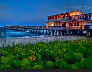 Un hotel frente a la playa con un muelle que se adentra en el océano, un letrero que dice 'L.W.B. Pier & General Store', y una persona caminando sobre la arena.