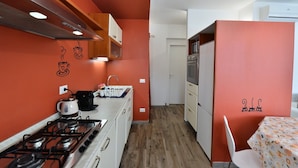 Een compacte keuken met een gasfornuis, een koffiezetapparaat en een magnetron. Er is een spoelbak met een kraan en kastjes erboven. De muren zijn oranje en er ligt een houten vloer. In de hoek staat een eettafel met een tafelkleed met dessin.