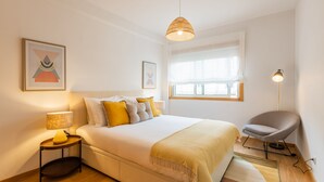A modern bedroom with a bed, bedside table, lamp, chair, and artwork on the walls. - Modern 2BR Apart w/ AC and Parking in City Center (Porto)