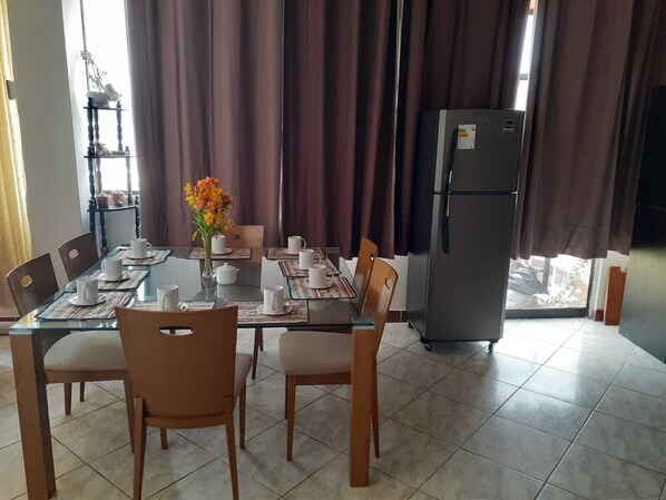 A dining area with a glass table, chairs, and a refrigerator.