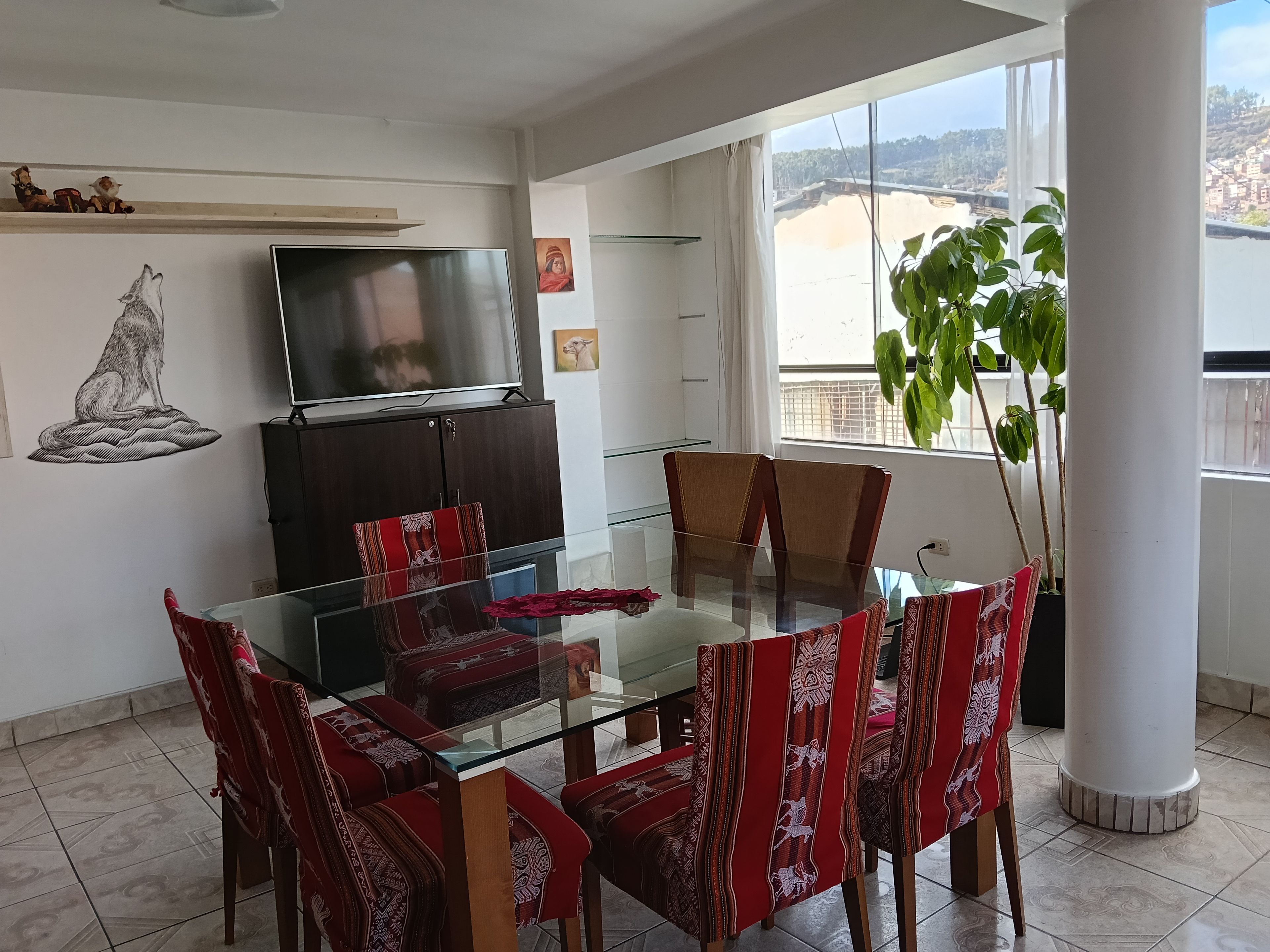 A dining area with a glass table and chairs, a television on a cabinet, and a view of a cityscape through large windows.