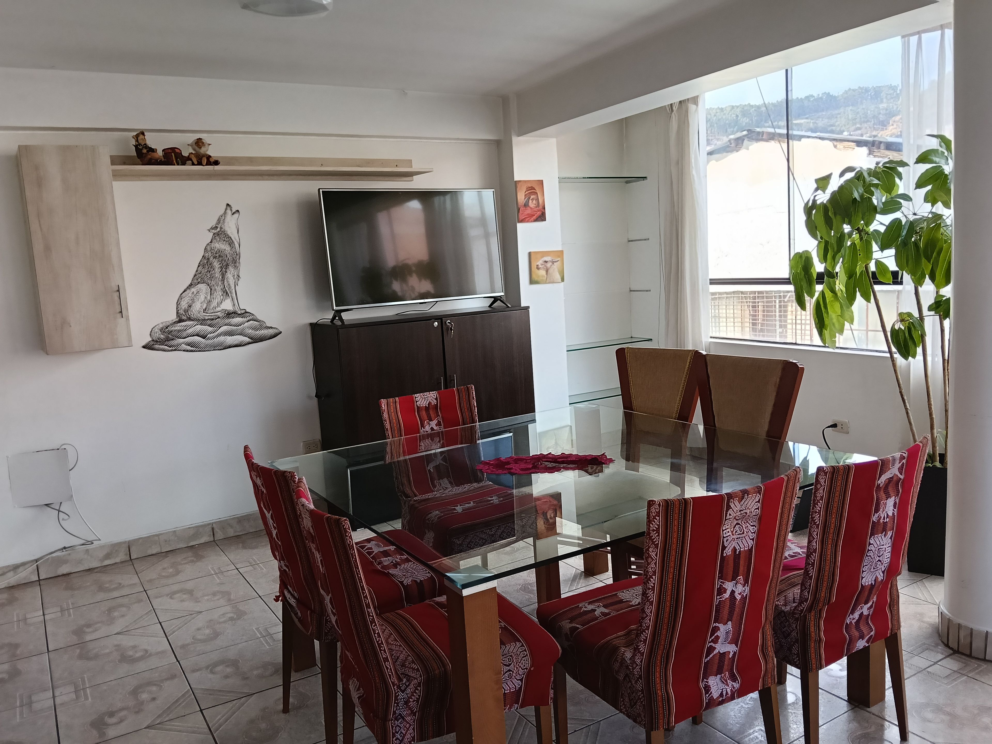 A dining area with a glass table and red patterned chairs, a dark wooden cabinet, and a television on the wall.