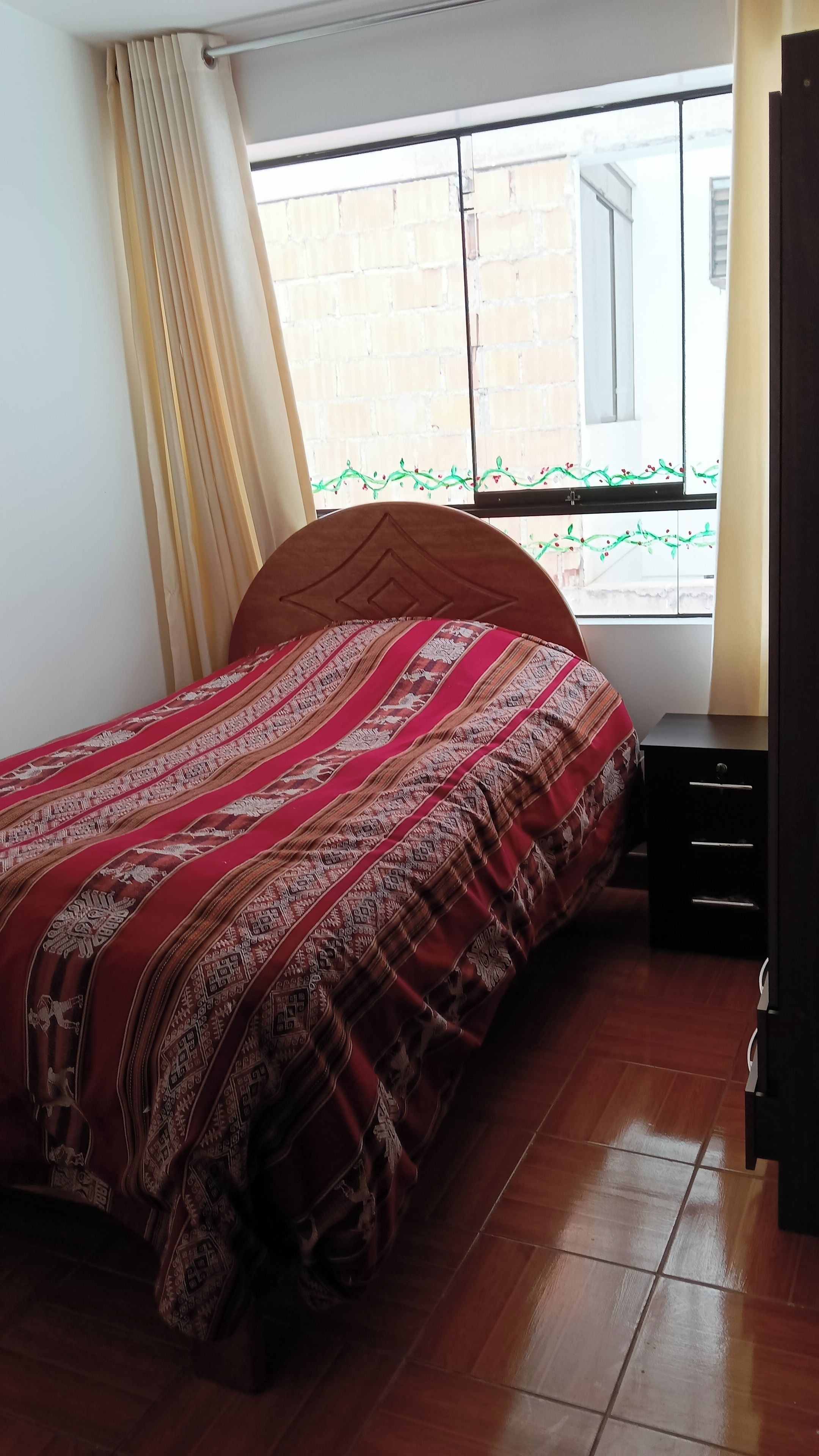 A bedroom with a wooden bed, a nightstand, a window with a view of a brick wall, and a door to the right.