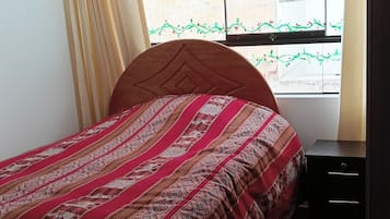 A bedroom with a wooden bed, a nightstand, a window with a view of a brick wall, and a door to the right.