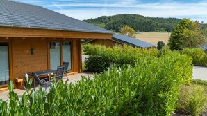 Eine Holzhütte mit einer Terrasse, großen Fenstern und Blick auf einen bewaldeten Hügel.