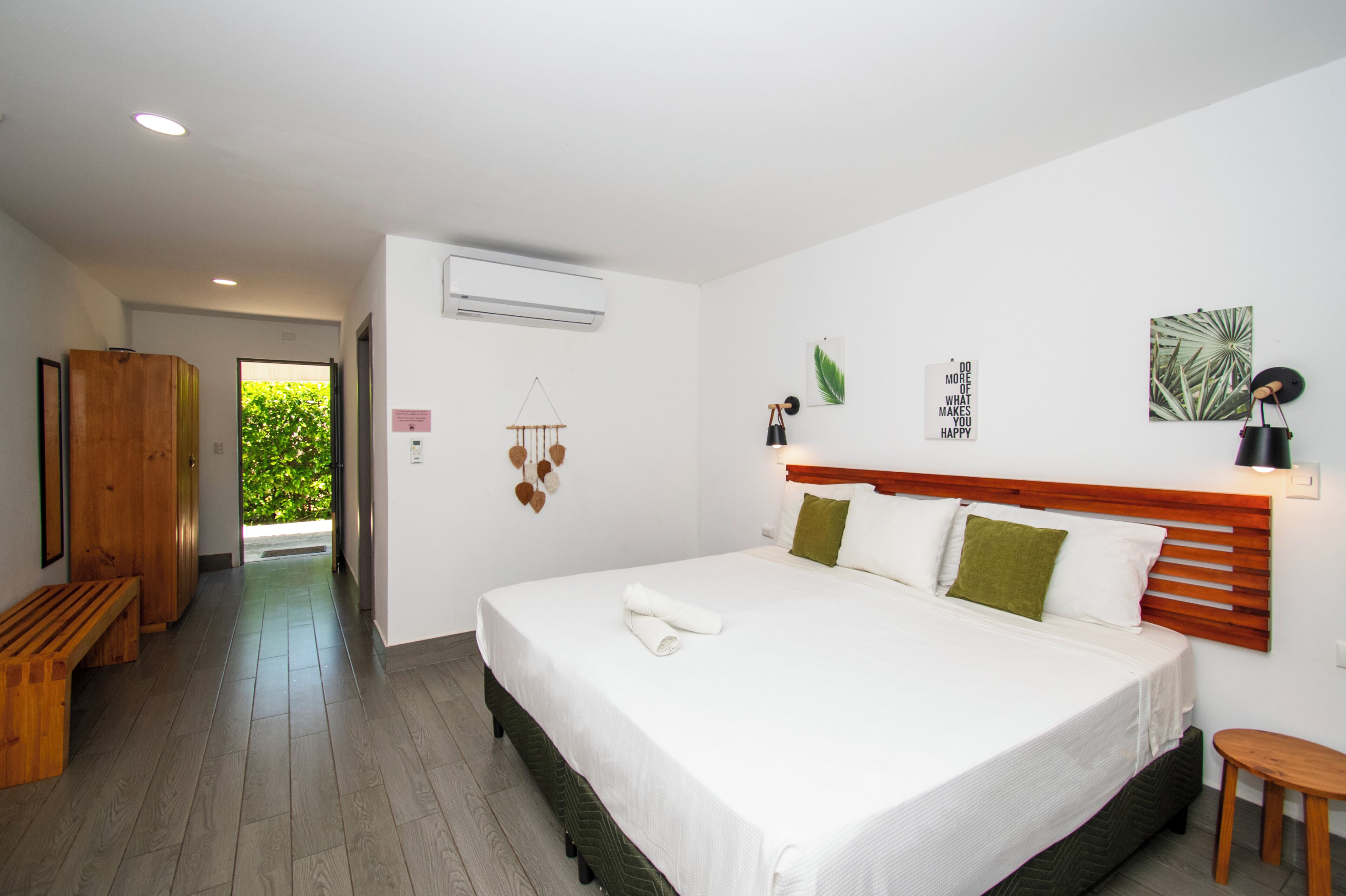 A hotel room with a large bed, a wooden bench, an air conditioning unit, and a view of greenery outside.