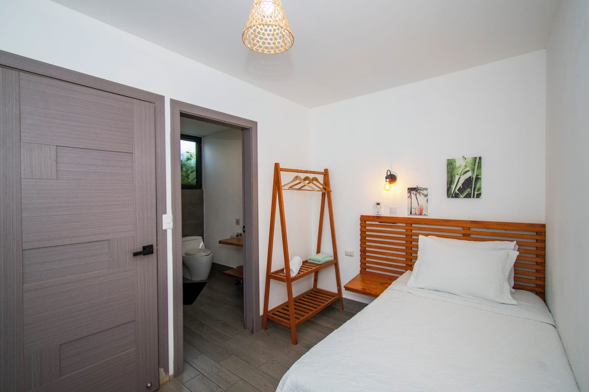 Un dormitorio con cama de madera, un perchero, una mesita de noche y una puerta que da a un baño.