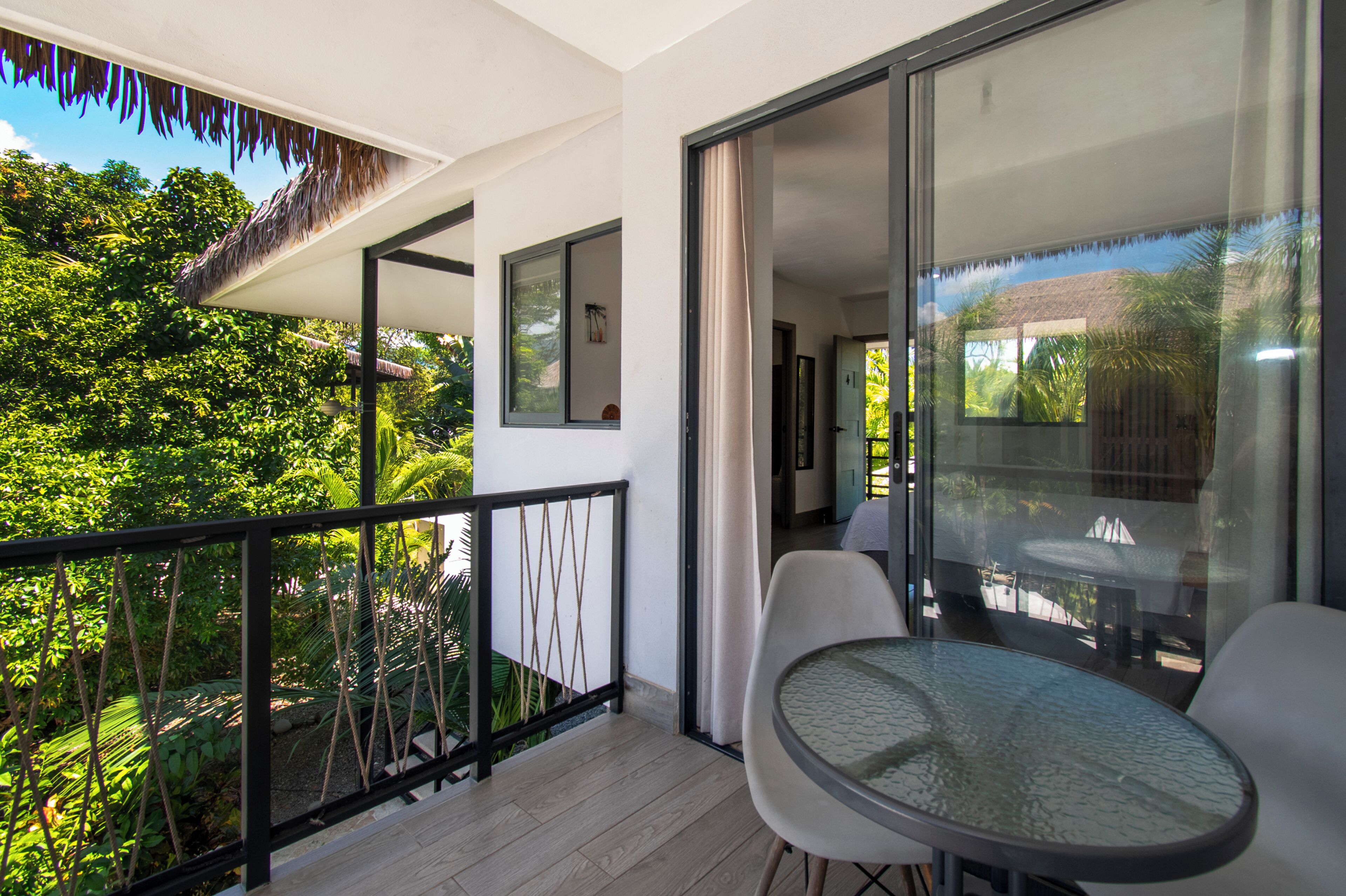 Un balcon avec une porte vitrée, une table ronde et des chaises, donnant sur un jardin luxuriant.