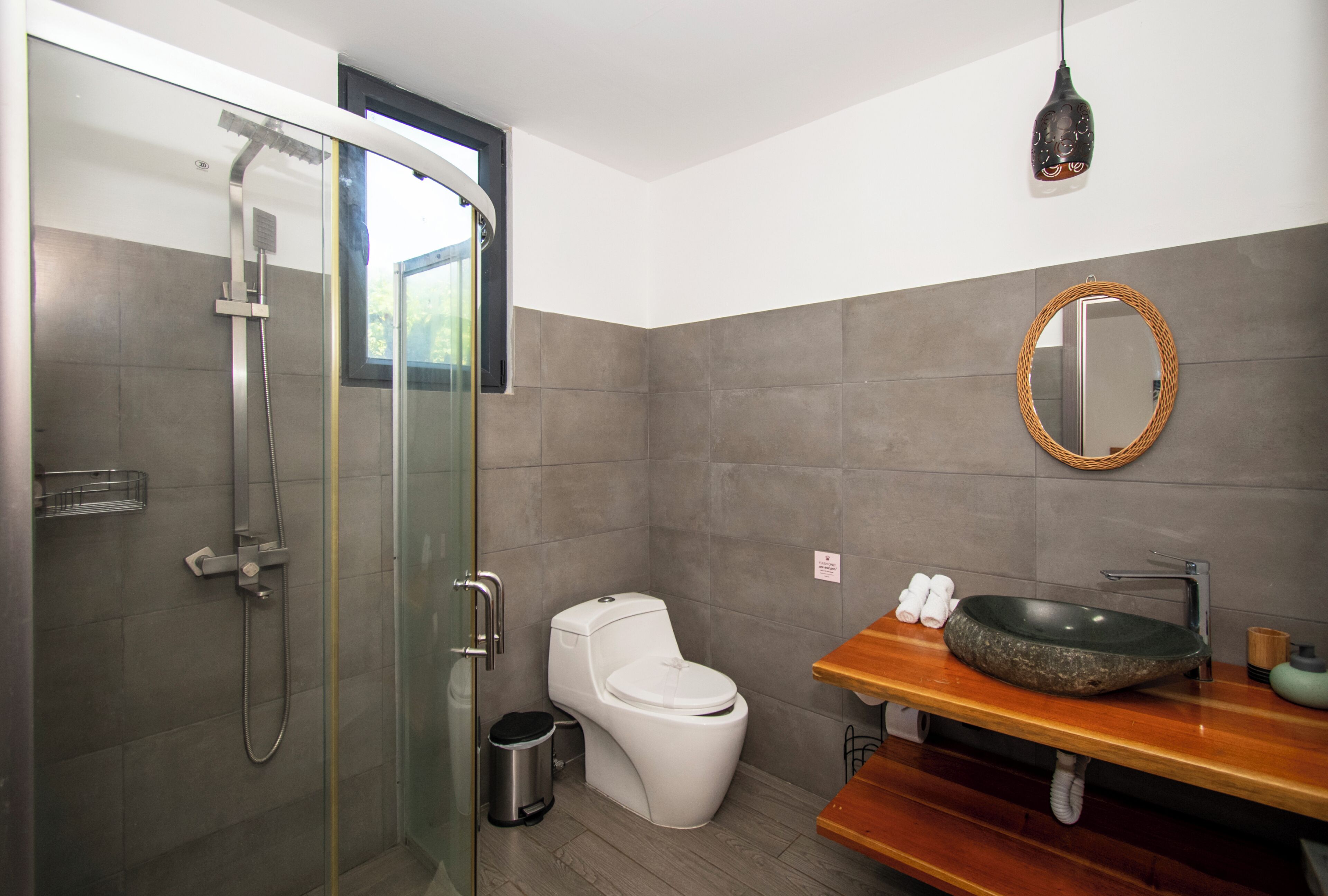 A modern bathroom with a glass-enclosed shower, a white toilet, and a wooden vanity with a round mirror.