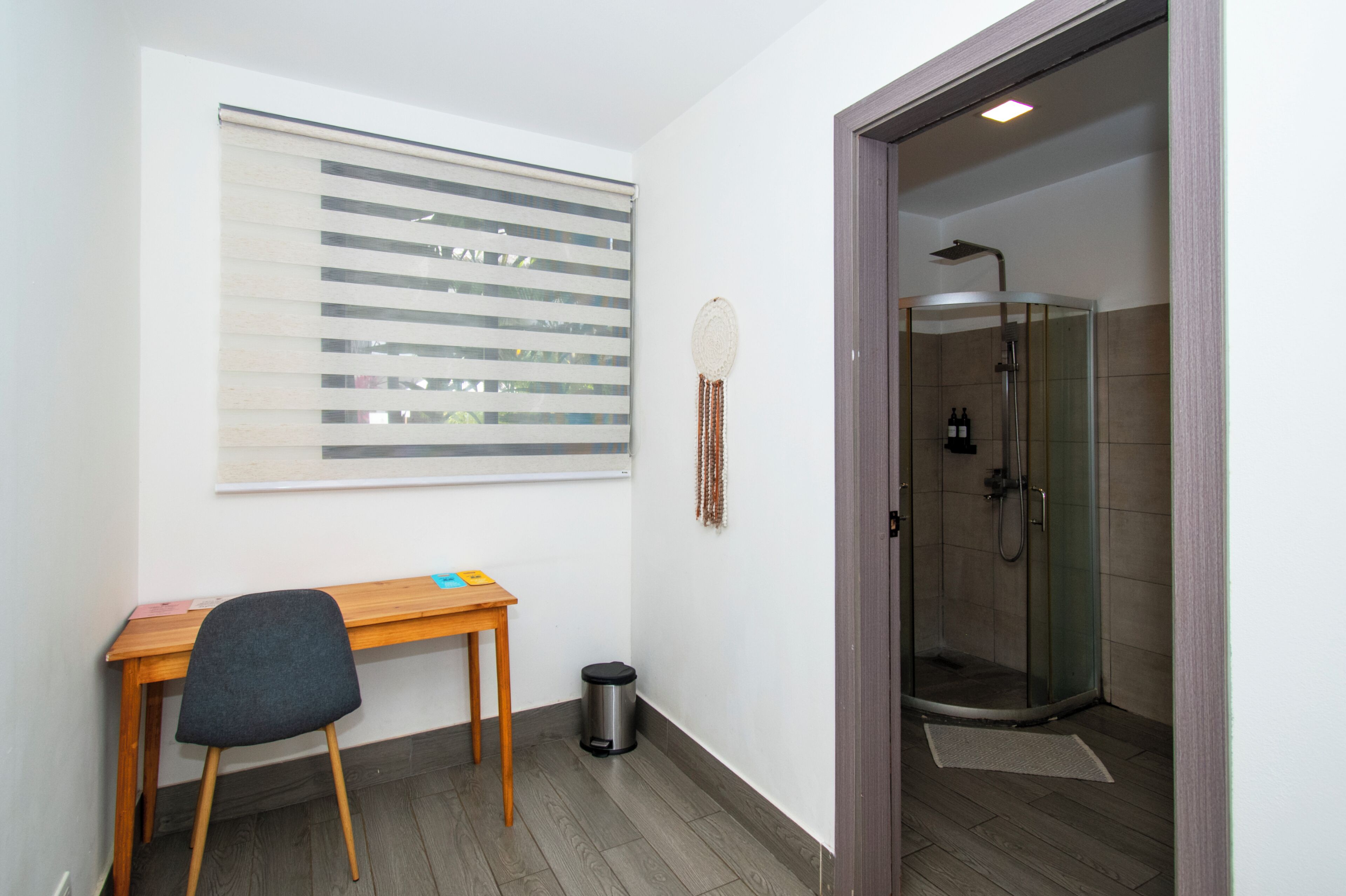 A room with a desk, chair, window with blinds, a decorative wall hanging, and a glass-enclosed shower area.