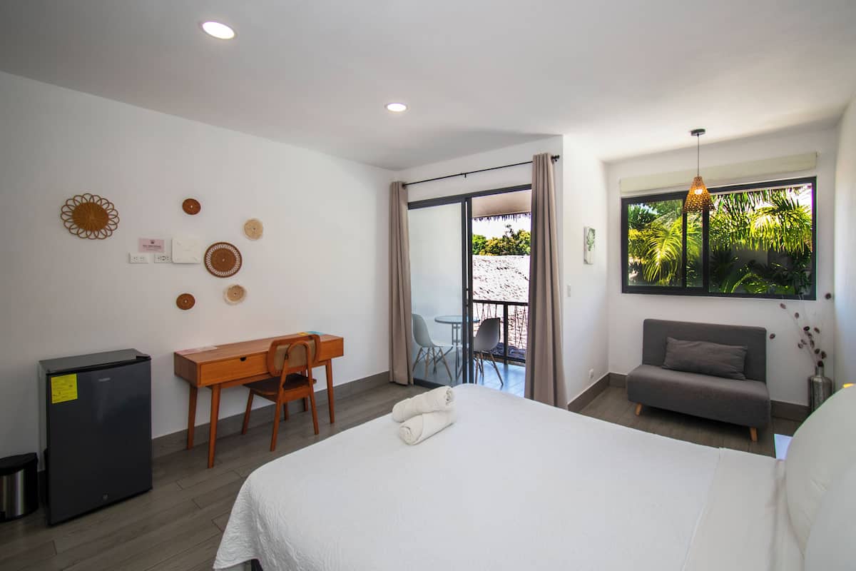 Habitación de hotel con cama, escritorio, silla, sofá, mesita, cesto de basura y ventana con vistas a la vegetación.
