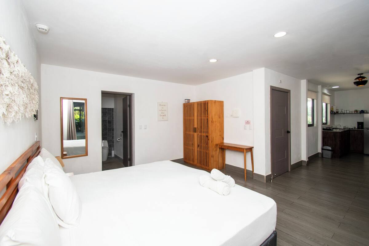 Una habitación de hotel moderna con una cama grande, un armario de madera, una mesita pequeña y vistas al área de la cocina.