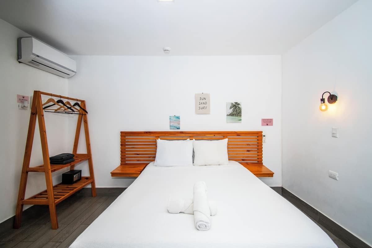 Una habitaci ón de hotel con una cama, un estante de madera, un aire acondicionado instalado en la pared y una lámpara fijada a la pared.