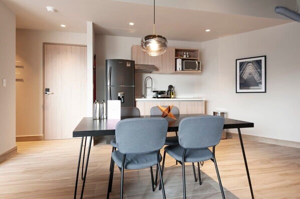 Cozinha moderna com frigorífico preto, micro-ondas e pia. Inclui mesa de jantar com duas cadeiras e uma luminária suspensa.