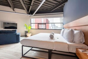 Quarto de hotel moderno com uma cama, um sofá azul, uma planta em vaso e uma grande janela com vista para a cidade.