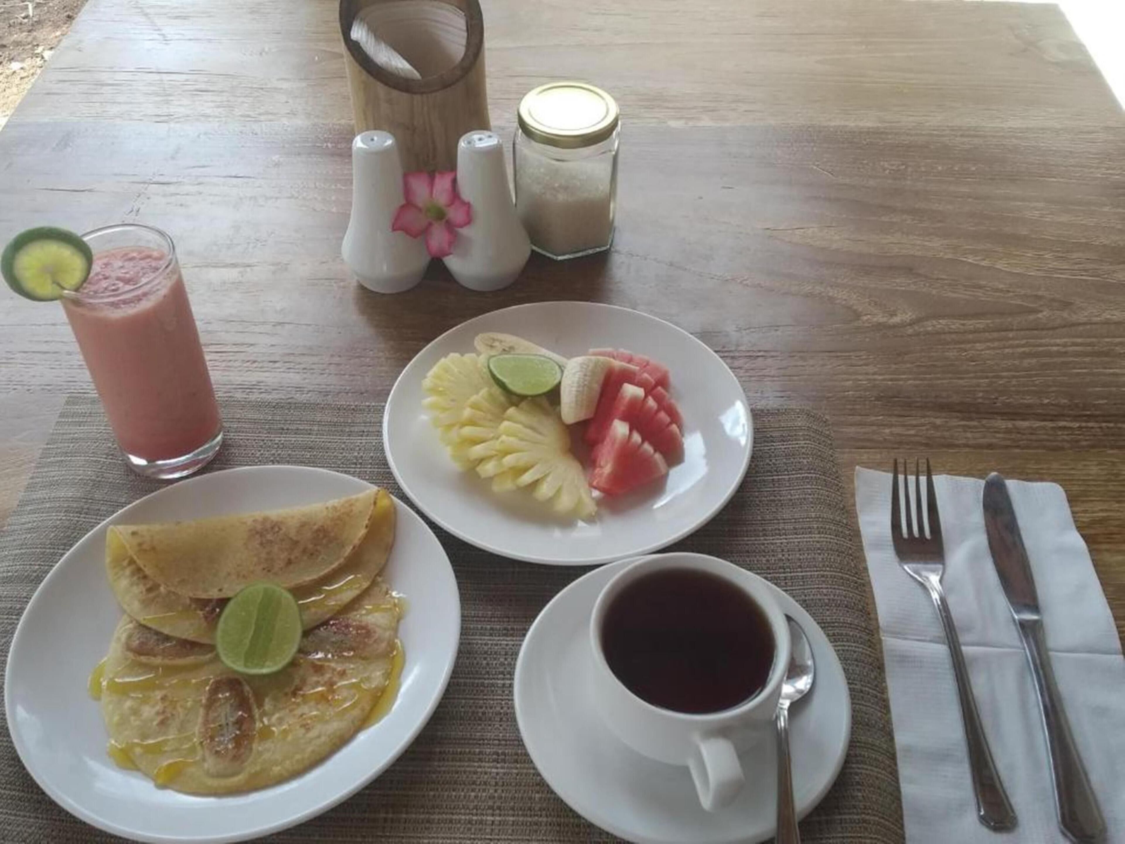 A breakfast table with a plate of pancakes, a plate of fruit, a glass of smoothie, and a cup of coffee.