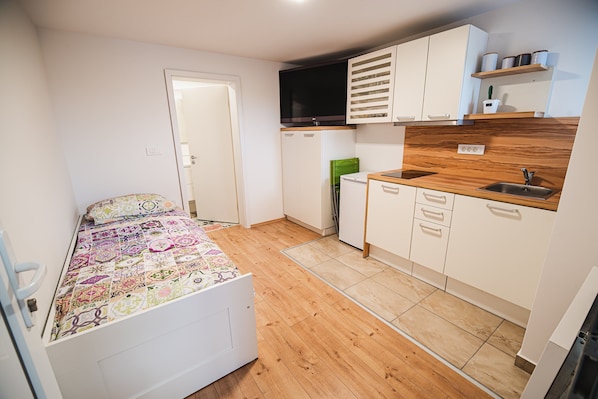 Quarto pequeno com cama, cozinha compacta e porta que dá acesso a outro espaço.