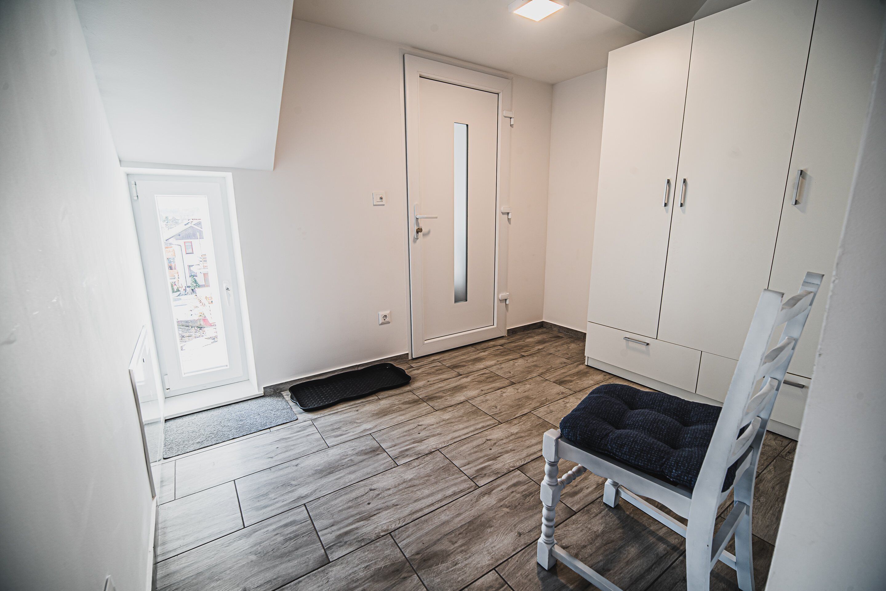 A room with a wooden floor, a white chair with a cushion, a white wardrobe, and a door with a view of the outside.