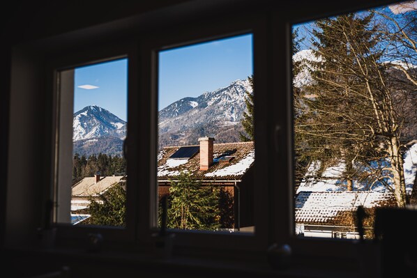 A view through a window of a mountain landscape with snow-capped peaks, a clear sky, and a building with a tiled roof.