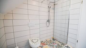 Une salle de bain avec douche, toilettes et carrelage au sol, le couvercle des toilettes étant décoré d’un motif en forme de cœur.
