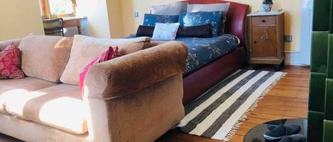 A cozy living room with a sofa, a bed with multiple pillows, a small wooden cabinet, and a round coffee table with shoes on it.
