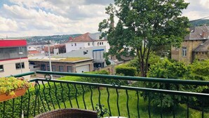 Un balcon avec une chaise en osier et une petite table, donnant sur un jardin verdoyant et sur des bâtiments en arrière-plan.