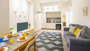 A modern living room with a grey sofa, a dining table with breakfast, and a kitchen in the background.