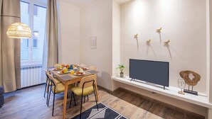 A modern living room with a dining table set for breakfast, a flat-screen TV mounted on the wall, and a large window with curtains.