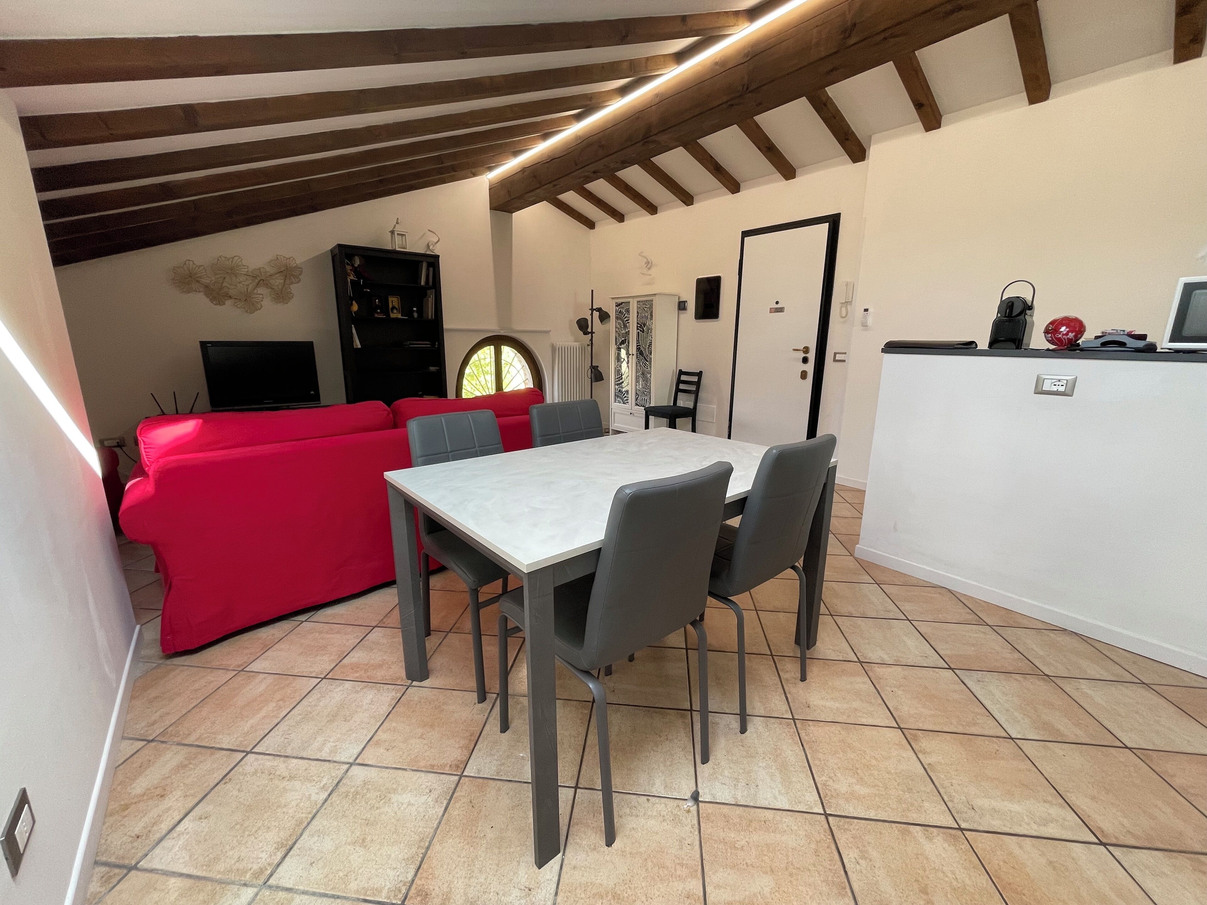 Zona pranzo con tavolo e sedie, un divano rosso e angolo cottura con piano di lavoro ed elettrodomestici.