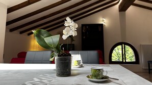 Una habitación con techo alto, vigas de madera, una mesa con una taza y un platillo, una planta en maceta y vistas a través de una ventana.