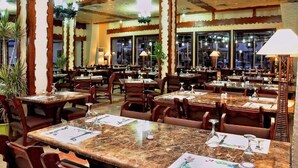 Ein Restaurantinneres mit Holztischen und -stühlen, großen Fenstern und hängenden Lampen.