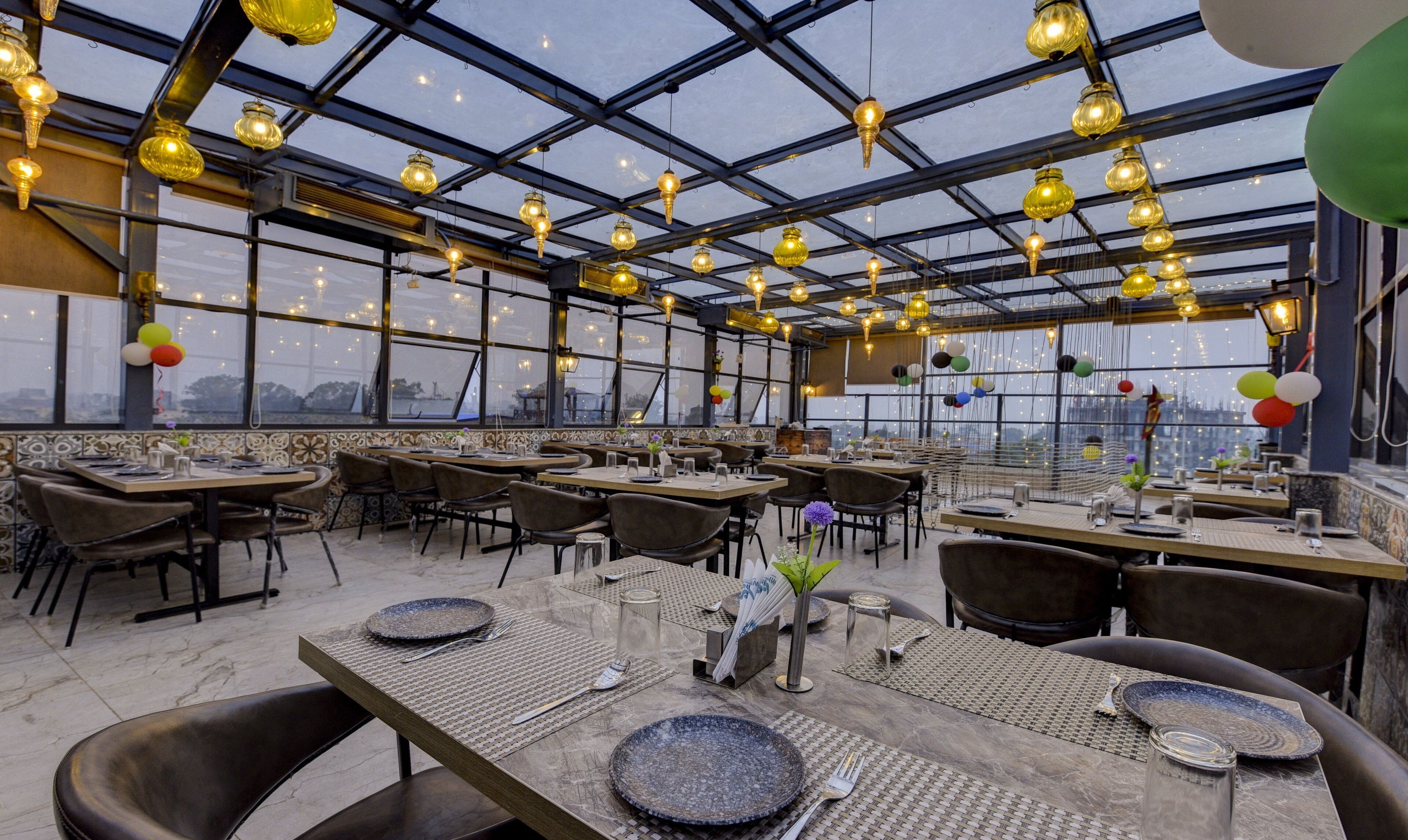 Un restaurante con techo alto y ventanas amplias, mesas dispuestas con platos y cubiertos, y lámparas colgantes.