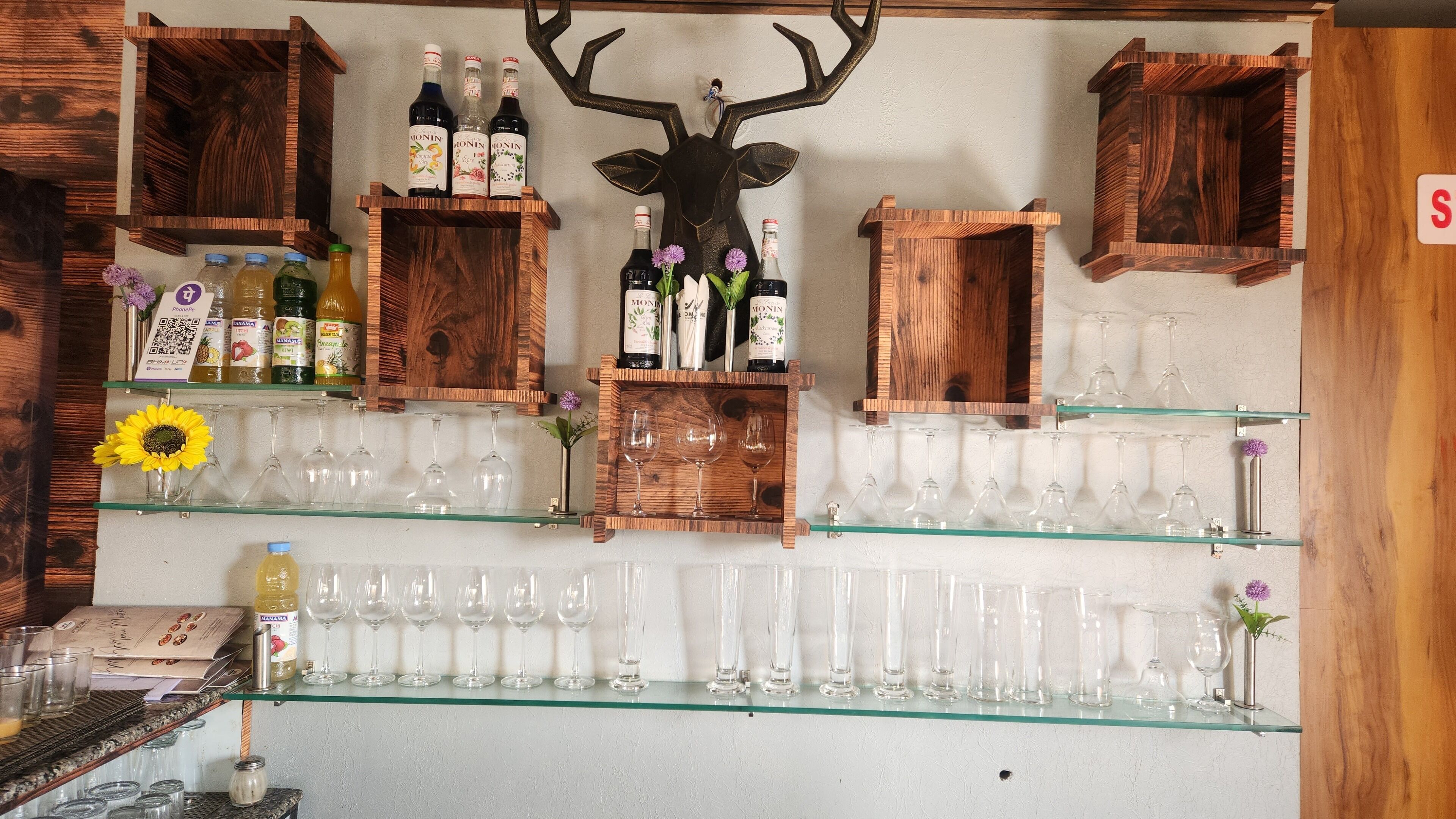 Un espace bar avec des étagères en bois où sont disposées des bouteilles et des verres, une tête de cerf servant de décoration, et un panneau avec un code QR.